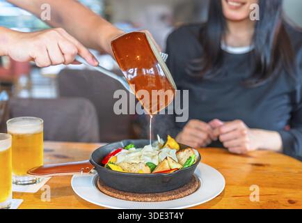 Konzentrieren Sie sich auf die Hände eines Kellners im Restaurant, der eine Keramikpfanne hält und geschmolzenen Käse auf eine gusseiserne Schüssel mit Brotcroutons und gießt Stockfoto