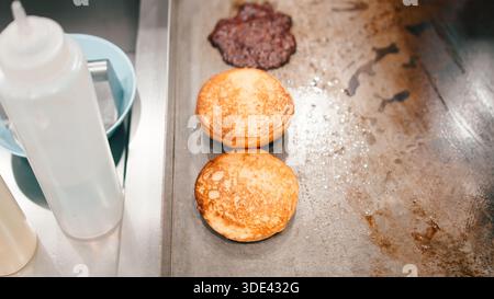 Burgerbrötchen, die zusammen mit einem Fleischpatty auf einem gewerblichen Grilltisch mit flacher Oberseite toasten Stockfoto
