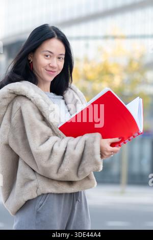 Eine junge Frau liest ein rotes Notizbuch vor dem Hintergrund von Stadtgebäuden Stockfoto