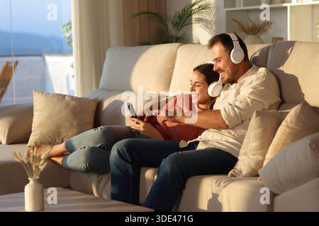 Glückliches Paar, das Kopfhörer trägt und auf einem Sofa sitzt, das zu Hause auf dem Telefon lauscht Stockfoto