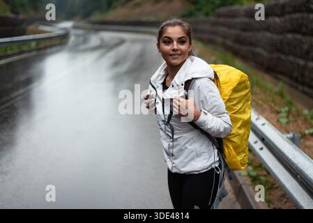 Eine junge Frau mit einem gelben Rucksack, die im Regen auf einer nassen Straße steht Stockfoto
