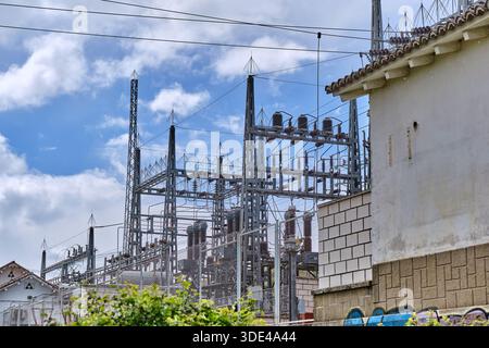 Stahlkonstruktionen einer elektrischen Unterstation stehen hoch vor dem Hintergrund flauschiger Wolken. Ein benachbartes Gebäude zeigt Anzeichen für städtische Entwicklung in Stockfoto