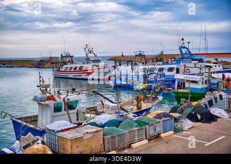 Fischerboote liegen an einem Hafen an. Sie sind ordentlich gebunden in ruhigem Wasser mit bewölktem Himmel darüber. Die Gegend zeigt Aktivitäten, die typisch für eine Küstenstadt sind. Stockfoto