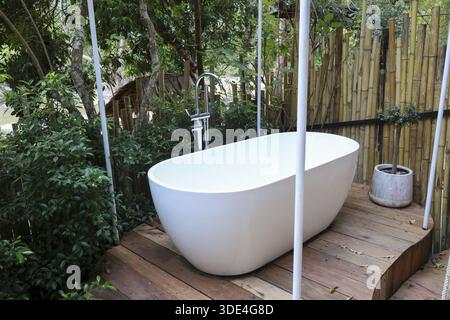 Ruhige weiße Badewanne im Freien auf Holzterrasse entspannender tropischer Garten Spa friedlicher Bambuszaun frische grüne Natur Luxus Ferienresort privates Bad Stockfoto