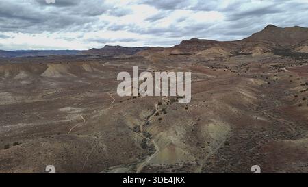 Aus der Vogelperspektive auf trockenen Badlands, die von gewundenen Pfaden unter einem stimmungsvollen Himmel gemeißelt wurden, stehen die gedämpften Erdtöne der Landschaft im Kontrast zu den weit entfernten, zerklüfteten Bergen Stockfoto