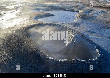 island kochender Schlammtopf auf eisigem Vulkanfeld, dampfender Kraterrand mit sprudelndem grauem Schlamm und glitzernder Mineralkruste, raue niedrige Sonne, die einen scharfen Schlamm hervorbringt Stockfoto