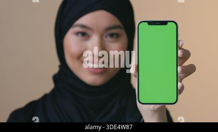 Lächelnde asiatische muslimische Frau in schwarzem Hijab, die Smartphone-Grünbildschirm hält und auf Kamera-Promo-Frau schaut, die Handy-islamisches Mädchen zeigt Stockfoto