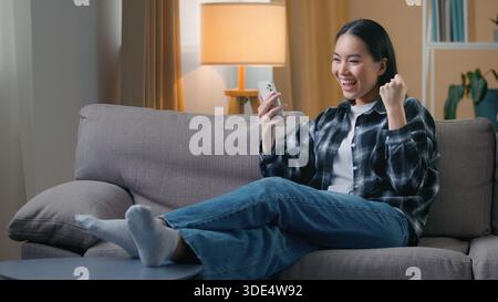 Glückliche asiatische Frau sitzend Sofa Haus feiert Sieg auf Smartphone fröhliche koreanische Frau spielt Handy Spiel gewinnen lesen gute Nachrichten Jobangebot Stockfoto