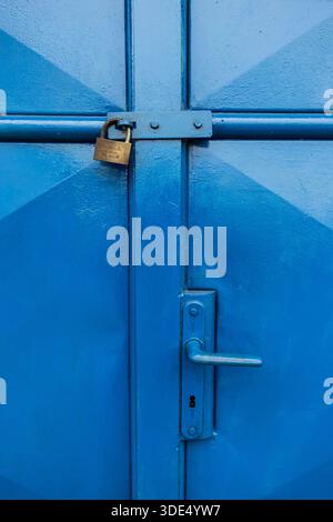 Nahaufnahme einer blauen Metalltür, die mit einem Vorhängeschloss gesichert ist und industrielle Texturen und verwitterte Farbe zeigt Stockfoto