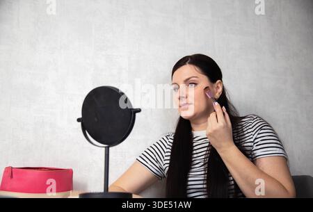 Erwachsene Frau, dunkles Haar und gestreiftes Hemd, trägt Gesichtsschminke mit Pinsel auf, während sie die Reflexion im Spiegel betrachtet. Rosafarbene Kosmetiktasche sichtbar auf Tisch o Stockfoto