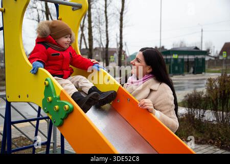 Junge in roter Winterjacke, braunem Hut, sitzt auf der gelben Spielplatzrutsche. Eine lächelnde Frau im beigefarbenen Mantel steht in der Nähe. Glückliche Interaktion. Outdoor Park, cool Stockfoto