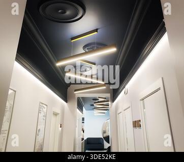 Moderne Inneneinrichtung des langen Flurs. Die dunkle Decke ist mit dekorativen Zierleisten ausgestattet. Lineare Pendelleuchten beleuchten den Raum und schaffen eine helle Atmosphäre. Anzeigen z. B. Stockfoto