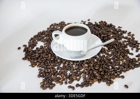 Eine Tasse Kaffee, die auf eine Oberfläche gelegt wird, auf der sich verstreute Kaffeebohnen befinden. Die Komposition unterstreicht die satten Brauntöne und das Aroma frischer Röste Stockfoto