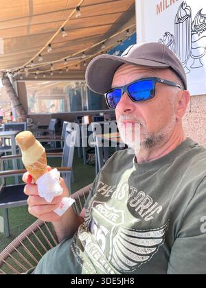 Älterer Mann mit blauer Spiegelsonnenbrille und Kappe, der an einem warmen Tag in teneriffa, spanien, Eisbecher genießt, während er in einem Café im Freien sitzt Stockfoto