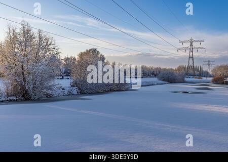 Winterlandschaft in den Niederlanden mit einem gefrorenen Kanal, schneebedeckten Bäumen und Hochspannungsmasten vor blauem Himmel. Stockfoto
