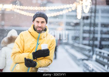 Mann mittleren Alters hält eine Tasse heißen Kaffee oder Tee auf der Eisbahn, im Freien an schneebedeckten Wintertagen Stockfoto