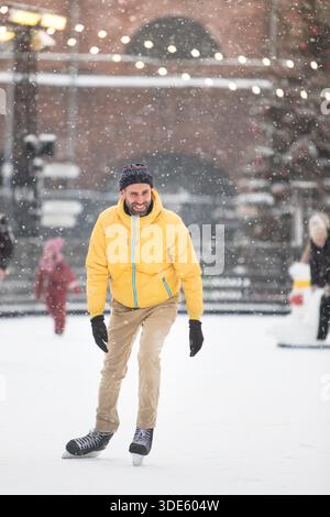 Fröhlicher Mann mittleren Alters in gelber Jacke, beigefarbene Hosen auf der Eisbahn, draußen an schneebedeckten Wintertagen Stockfoto