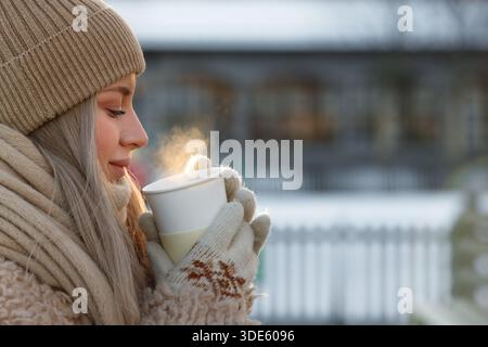 Zufriedene Frau hält im Winter draußen eine Tasse heißen Tee oder Kaffee, heißer Dampf kommt aus der Tasse Stockfoto