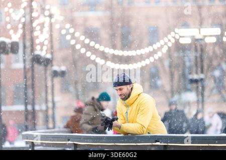Mann mittleren Alters, der in der Nähe des Zauns auf der Eislaufbahn steht und ein Smartphone benutzt, draußen an schneereichen Tagen Stockfoto
