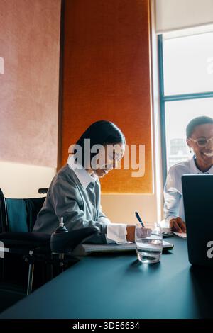 Zwei junge Profis, einer sitzt im Rollstuhl, tauschen sich während eines Arbeitstreffens in einer modernen Büroumgebung Ideen aus und zeigen Inklusivität und Tee Stockfoto