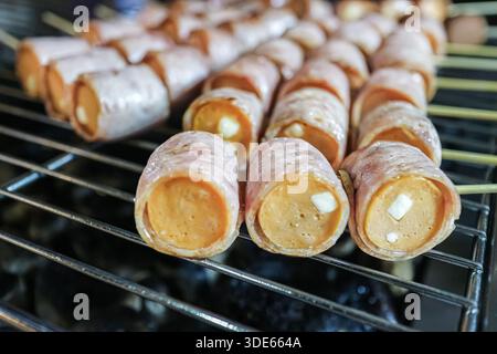 Mit Speck umwickelte Käsewürste auf einem Holzkohlegrill, die moderne thailändische Street Food Fusion und eine köstliche Snackkultur repräsentieren. Stockfoto