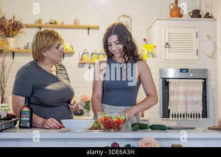 Zwei Frauen verschiedener Generationen kochen zusammen in einer gemütlichen Küche. Mutter und Tochter kochen zusammen in einer warmen Küche. Salat zubereiten Stockfoto