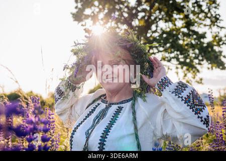 Eine Frau im Alter von 50 Jahren mit traditionellem ukrainischem besticktem Hemd und Blumenkranz steht bei Sonnenuntergang in einem blühenden Lupinenfeld. Kulturerbe Stockfoto