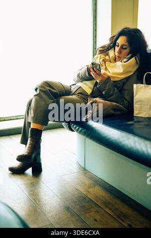 Die Frau entspannt sich, indem sie ihr Smartphone im Erfrischungsraum eines Schiffes ansieht. Die Frau trägt einen beigefarbenen Mantel, eine Hose und braune Stiefel Stockfoto