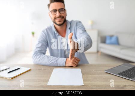 Konzept für Bewerbungsgespräche. Positiver Millennial-Mann, der die Hand zum Händeschütteln streckt, am Arbeitsplatz sitzt, selektive Konzentration Stockfoto