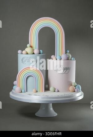 Eine kreative gemeinsame Geburtstagskuchen-Ausstellung mit zwei separaten Ebenen, silbernen Schriftnamen und einer hohen, pastellfarbenen Regenbogenbrücke, präsentiert auf einem weißen Sockel Stockfoto