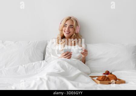 Junge Frau isst Frühstück im Bett hört Musik, genießt Freizeit, Wochenenden und Urlaub Stockfoto