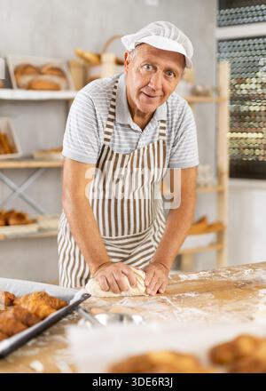 Senior Mann arbeitet in der Bäckerei als Bäcker, Knete Teig, arbeitet mit Mehl. Stockfoto