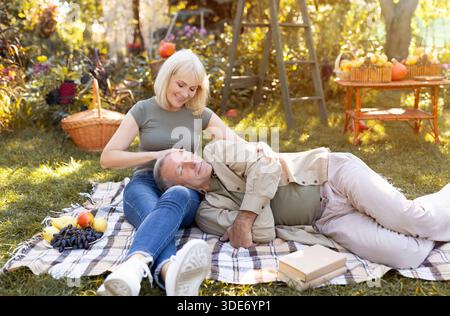 Ein zartes Seniorenpaar genießt die gemeinsame Zeit, ruht sich auf einer Picknickdecke im Garten aus und plaudert, Mann liegt auf den Beinen der Frau Stockfoto