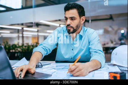 Der Techniker arbeitet tagsüber in der Büroumgebung an Projektplänen, während er Laptop und Stift verwendet Stockfoto