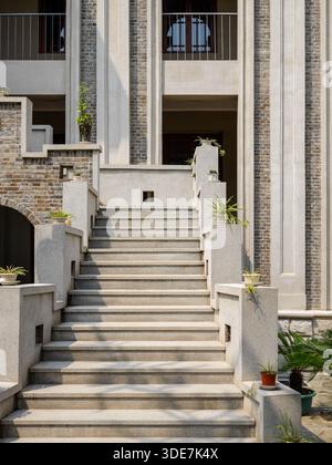 Architektonische Details einer Steintreppe, die zu einem Gebäudeeingang führt. Das Gebäude besteht aus grauen Ziegeln, Säulen und grünen Topfpflanzen. Die Stockfoto
