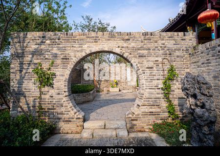 Klassisches kreisförmiges Mondtor in einer grauen Backsteinmauer, das zu einem traditionellen chinesischen Garten im Innenhof führt. Das Bild vermittelt kulturelles Erbe, Feng Shui und Stockfoto