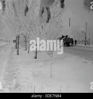 Slawjansk, Region Donezk, Ukraine, UdSSR. Winterfoto der frühen 1950er Jahre. Eine verschneite Straßenszene in der Stadt mit jungen Bäumen, die stark mit weißem Raureif bedeckt sind. Ein sowjetischer Vintage-Lastwagen, wahrscheinlich ein GAZ-Modell, transportiert eine Gruppe von Arbeitern im offenen Rücken entlang der vereisten Straße. Eine Frau im dunklen Mantel steht in der Nähe. Das Bild wird von verschwommenen, frostigen Zweigen im Vordergrund eingerahmt, die authentisch die raue, aber schöne Winteratmosphäre und die tägliche Arbeit im friedlichen Donbass während der Jahre der industriellen Erholung der Nachkriegszeit einfangen. Stockfoto