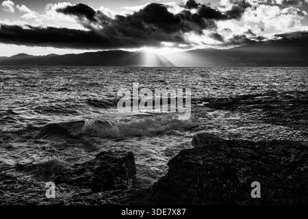 Schwarz-weiß-Ansicht von Wellen, die gegen Felsen im Vordergrund brechen, während Sonnenaufgangslicht unter schwerer Wolke über den Bergen von Carneddau strömt Stockfoto