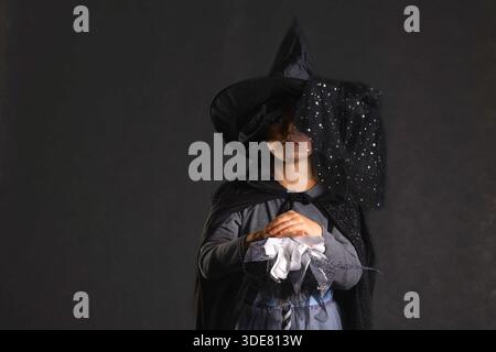 Mädchen in klassischem Hexenkostüm mit spitzem Hut und Schleier stehend mit gefalteten Händen im dunklen Studio Stockfoto