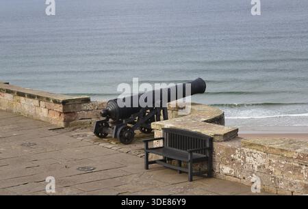 Bamburgh Castle in England : historische schwarze Kanone auf einer Steinplattform mit Blick auf die ruhigen Meereswellen und den Sandstrand mit einer Holzbank in der Nähe Stockfoto