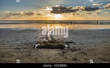 Eine ruhige Strandszene bei Sonnenuntergang mit Silhouetten einer kleinen Gruppe, die entlang des nassen Sandes schlendert, mit Sonnenstrahlen auf dem Wasser und einer warmen, friedlichen Atmosphäre Stockfoto
