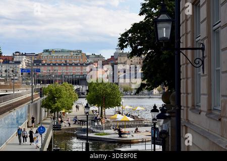 Im Sommer verbringen die Menschen Zeit an einer Uferpromenade in einer Stadt mit Fußgängern, Sitzplätzen im Freien und städtischen Gebäuden im Hintergrund. Stockfoto