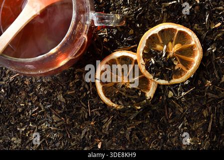 Details einer Glasschale mit heißem Schwarztee-Aufguss und Zitronenscheiben auf einem Hintergrund getrockneter Schwarzteeblätter. Draufsicht. Stockfoto
