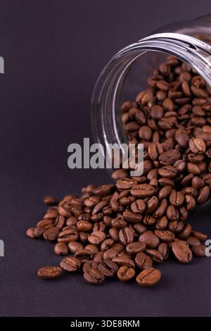 Geröstete Kaffeebohnen, die aus einem Glas verschüttet wurden. Dunkles Stillleben Stockfoto