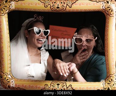 Eine Frau mit weißem Kleid und Sonnenbrille posiert für ein Foto. Eine Frau in einem blauen Hemd steht hinter ihr Stockfoto