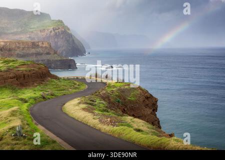 Kurvige Küstenstraße führt entlang dramatischer Klippen auf Madeira, Portugal, mit einem weichen Regenbogen, der nach Regen über dem Atlantik erscheint. Stockfoto