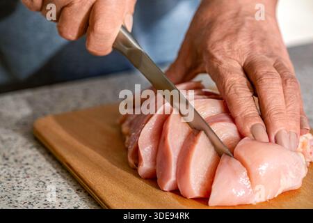 Koch-Hände schneiden das Hähnchenfilet mit einem Messer. Hähnchenschneidtechnologie. Man schneidet Hähnchenfilet auf Holzbrett, Tisch, Nahaufnahme. hef Woma Stockfoto