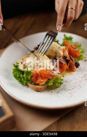 Eine Person genießt einen Brunch-Teller mit geräuchertem Lachs auf Toast, garniert mit pochierten Eiern und umgeben von frischem Gemüse. Gabel und Messer sind in Aktion Stockfoto