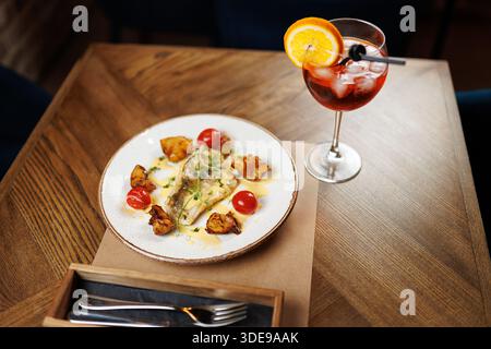 Der Esstisch ist mit gegrilltem Fisch, Kirschtomaten und frittiertem Gemüse ausgestattet. Ein farbenfroher Cocktail garniert mit einer Orangenscheibe sitzt besi Stockfoto