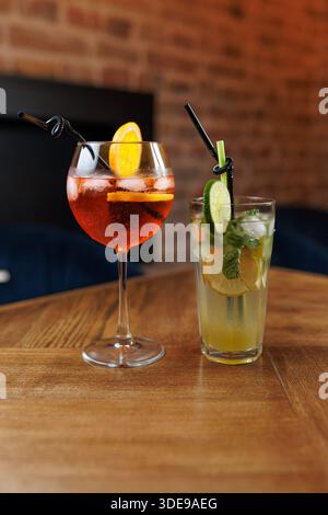 Zwei Cocktails sitzen auf einem Holztisch in einer Bar. Das eine ist ein helles Getränk mit Zitronenscheiben, das andere mit Pfefferminze und Gurke. Die Kulisse hat einen Krieg Stockfoto